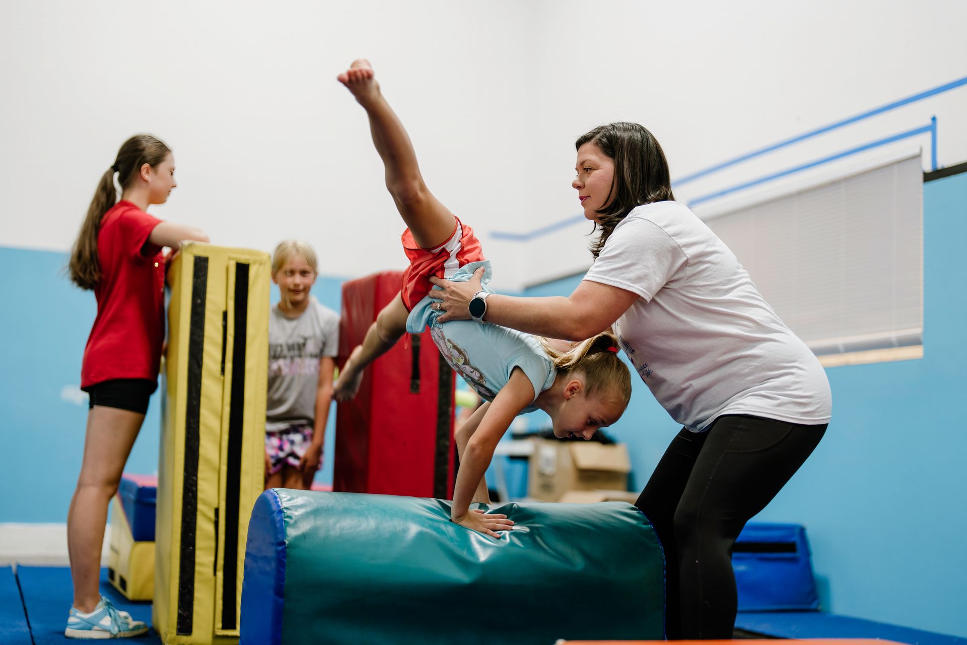 Gymnastics class with a child balancing on a padded vault while an instructor spots her.