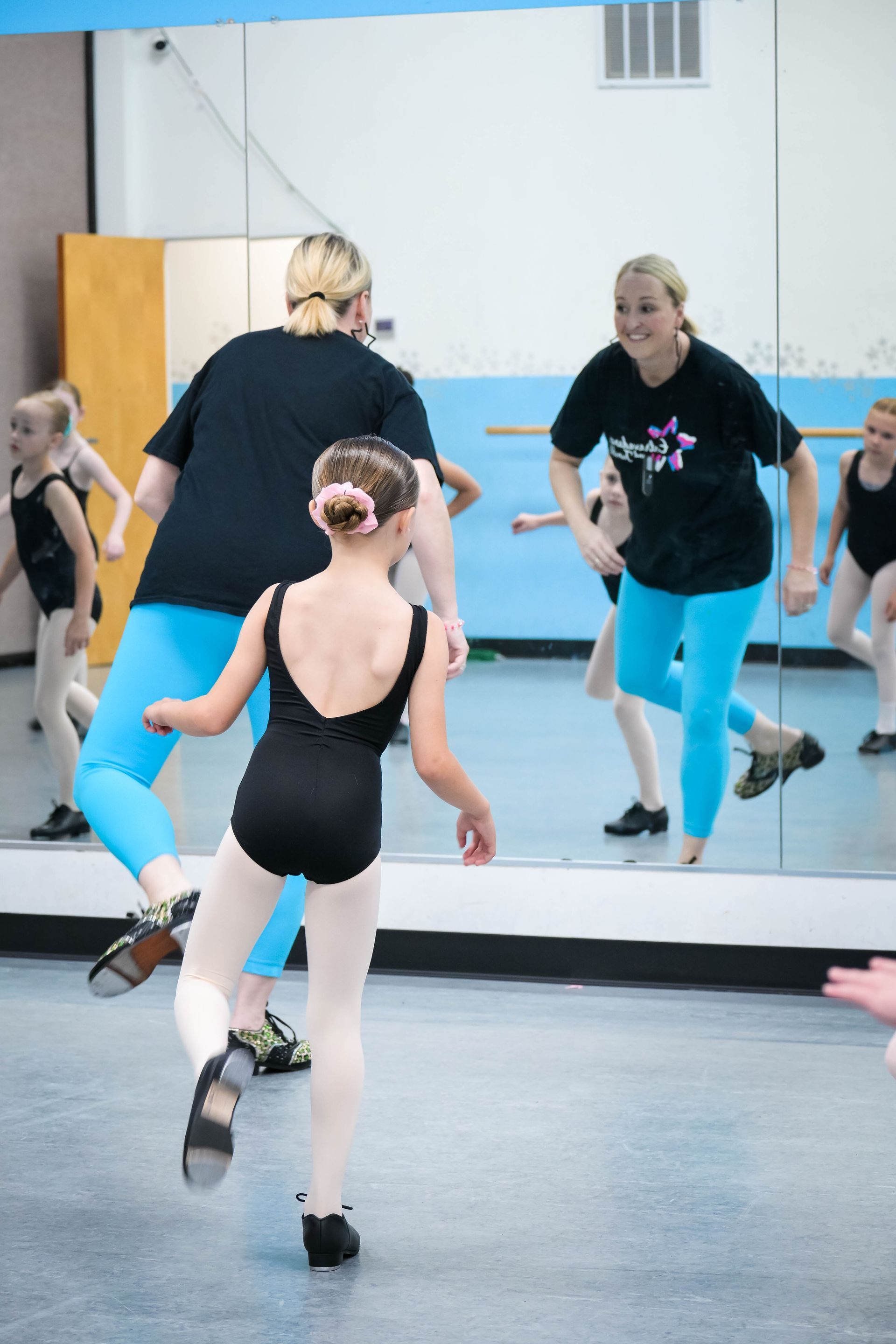 Dancers practicing in a studio, seen in a mirror with one dancer in a black leotard and bright blue leggings.
