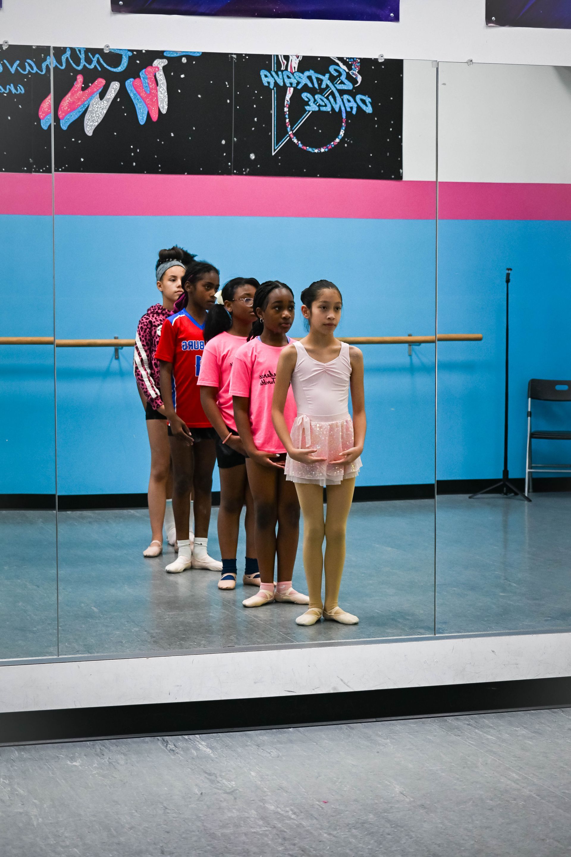 Five dancers stand in line in a studio, with the front dancer in pink and tan on a mirrored floor.