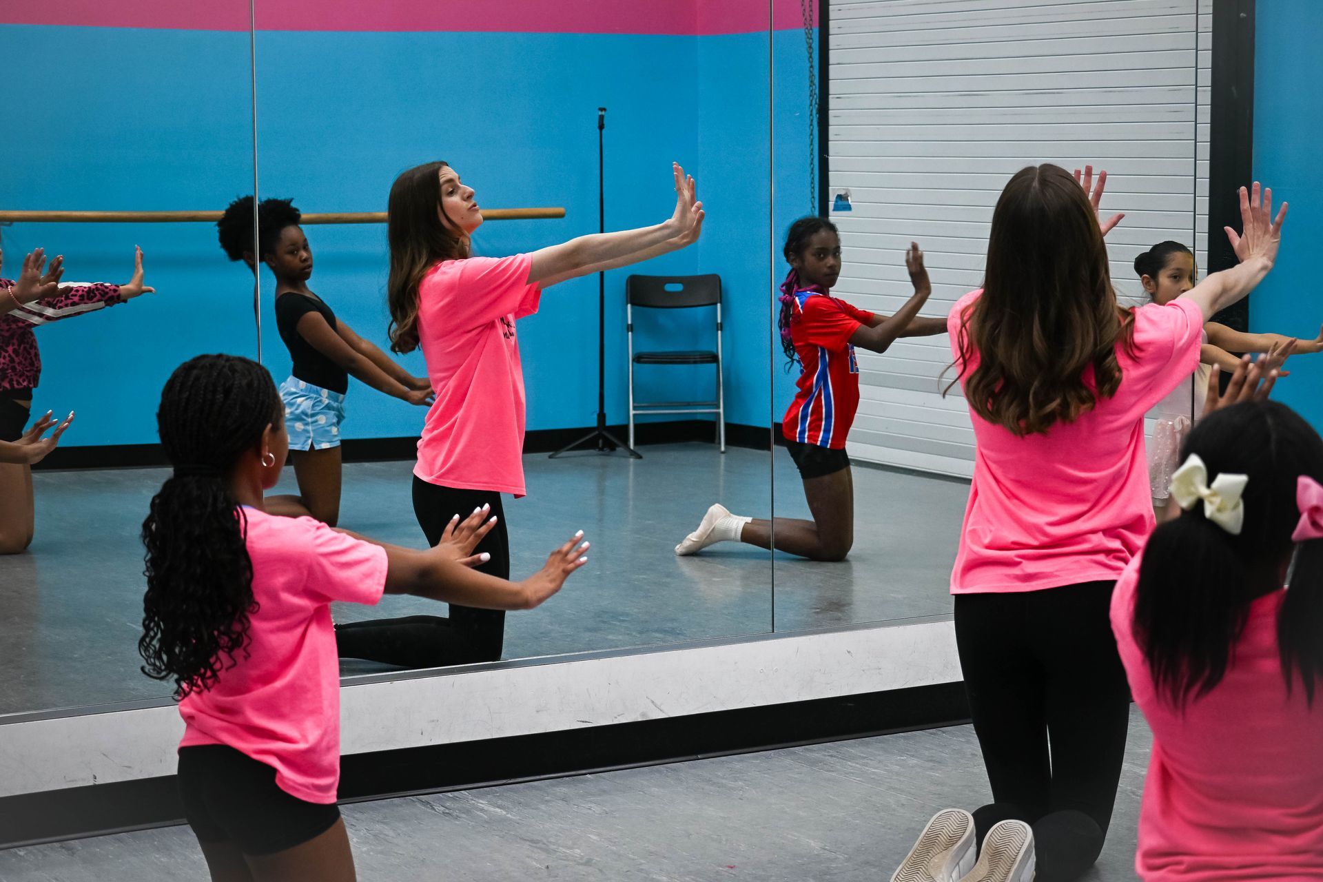 Dance class in a mirrored studio, students practicing arm movements in pink and black workout clothes