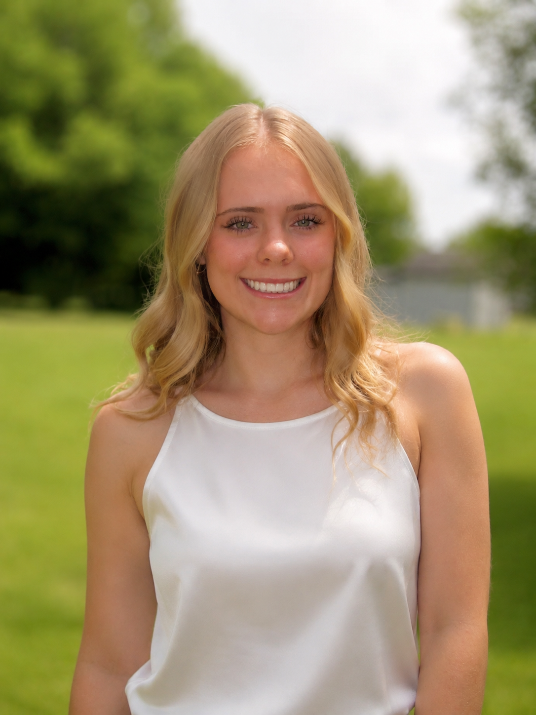 Smiling blonde woman in a white sleeveless top standing outdoors with green trees behind her