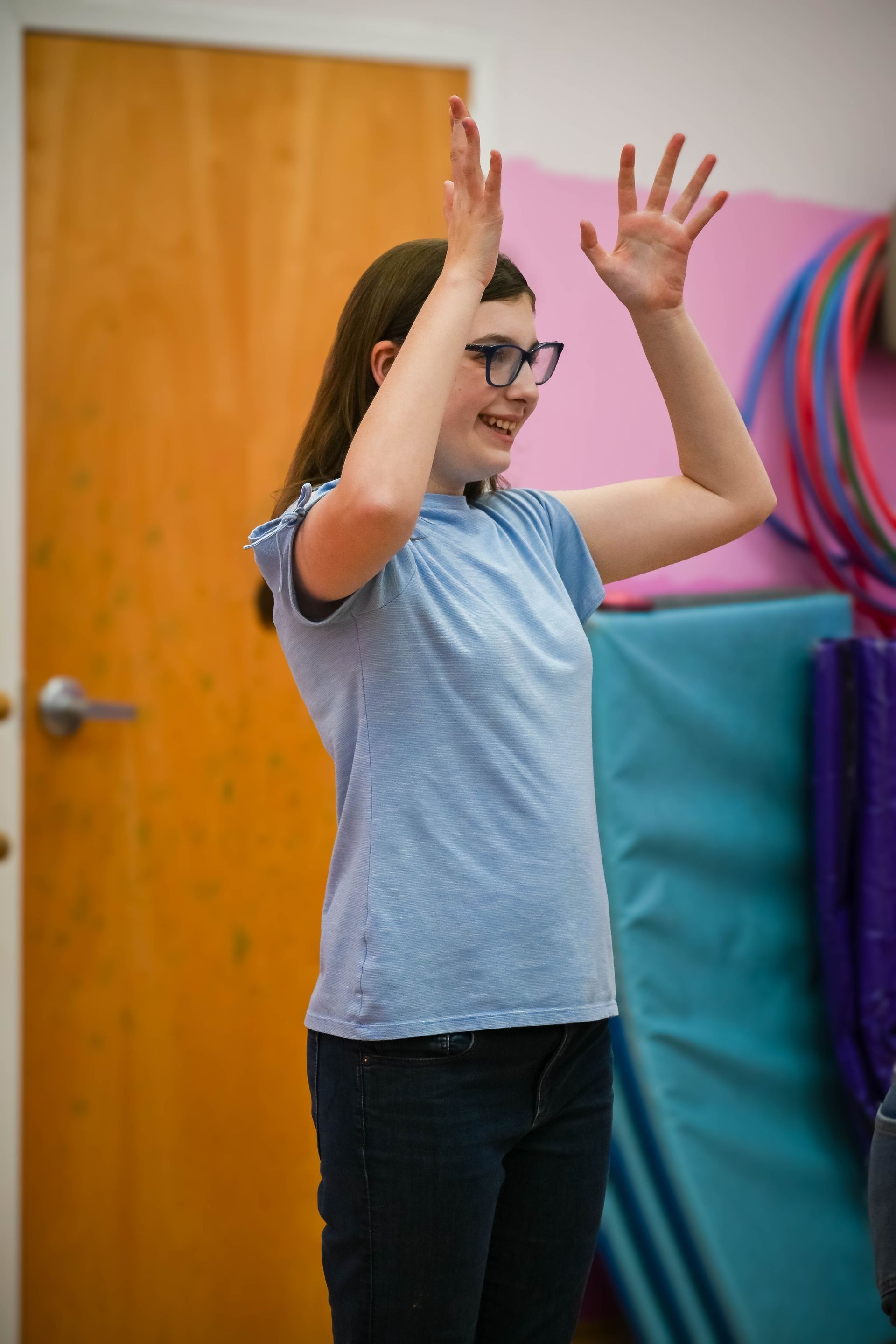 Child raising hands in a classroom, wearing glasses and a blue shirt, with colorful play mats in the background