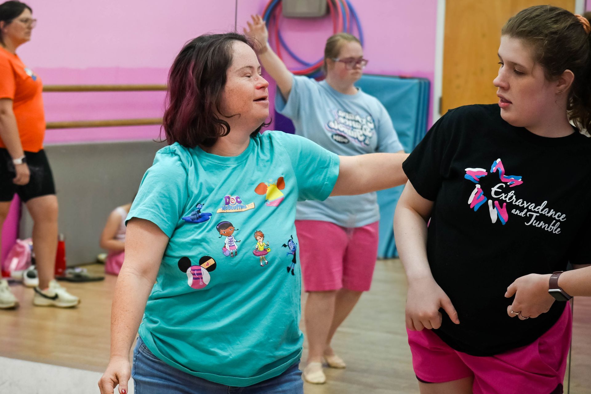 Two women dancing in a gym, one in a teal shirt reaching out to the other in a black shirt and pink shorts.
