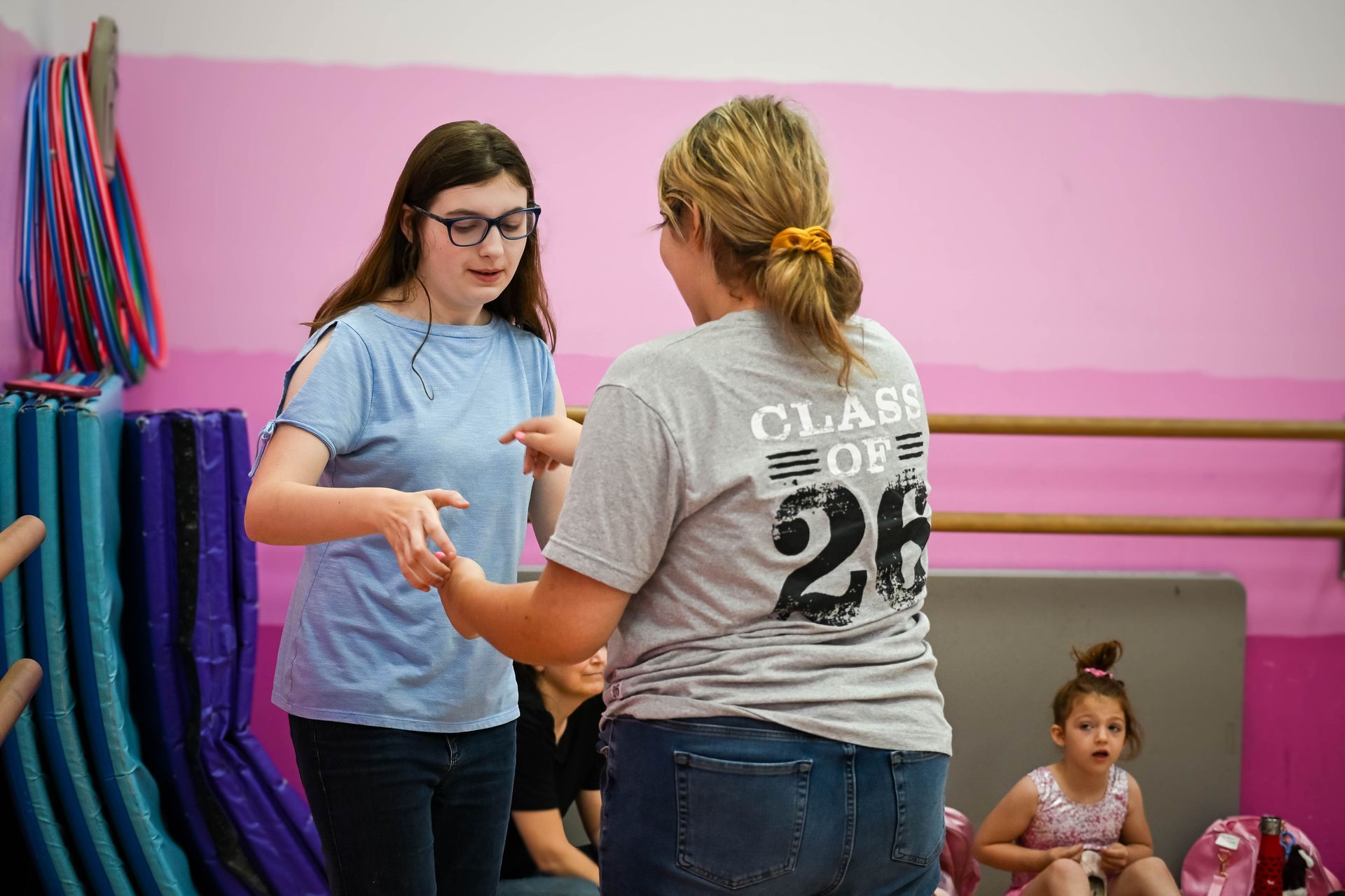 Two women in a gym adjusting equipment, with mats and colorful straps against a pink wall.