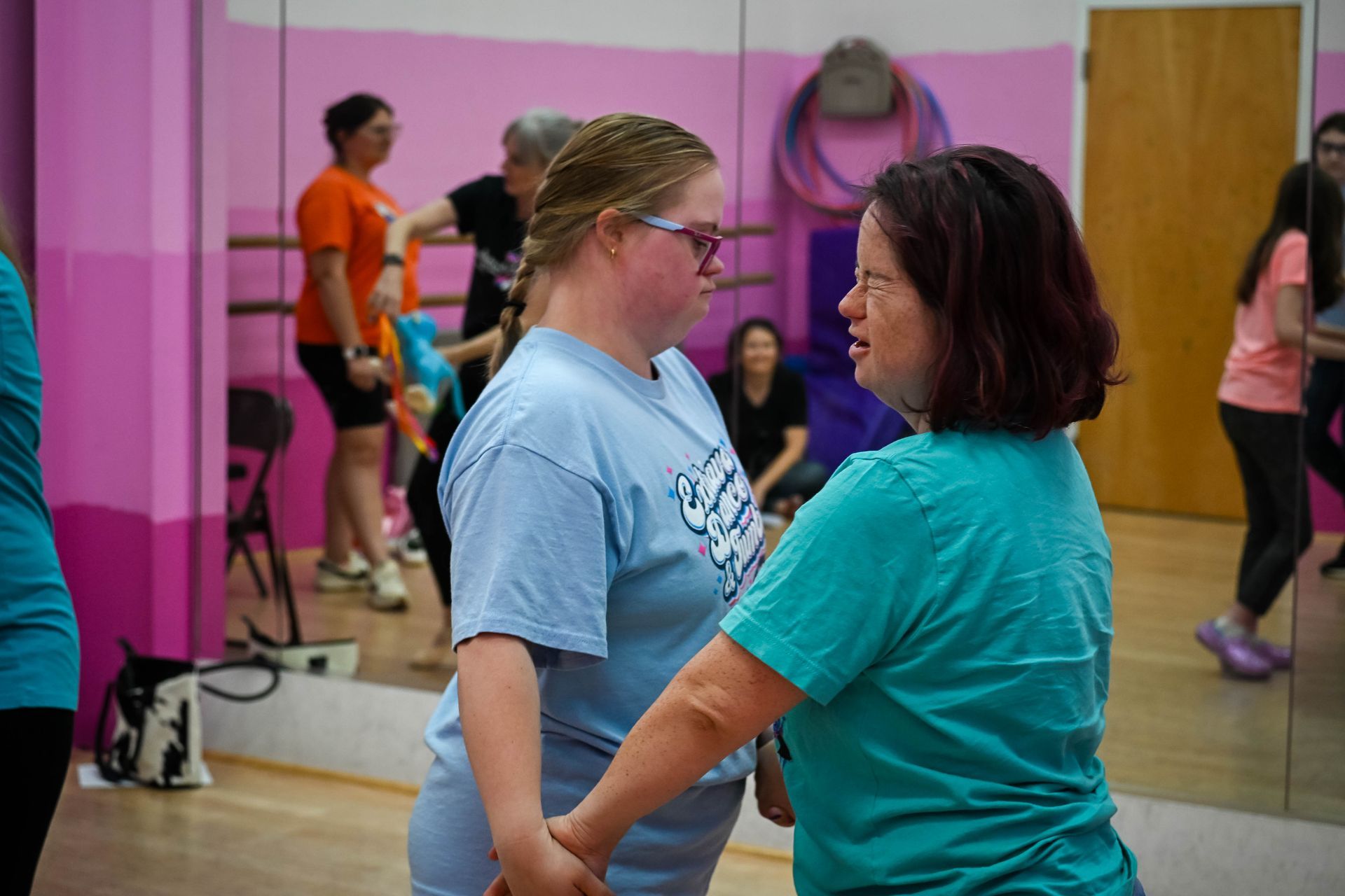 Two people holding hands in a mirrored dance studio with pink walls and other dancers in the background