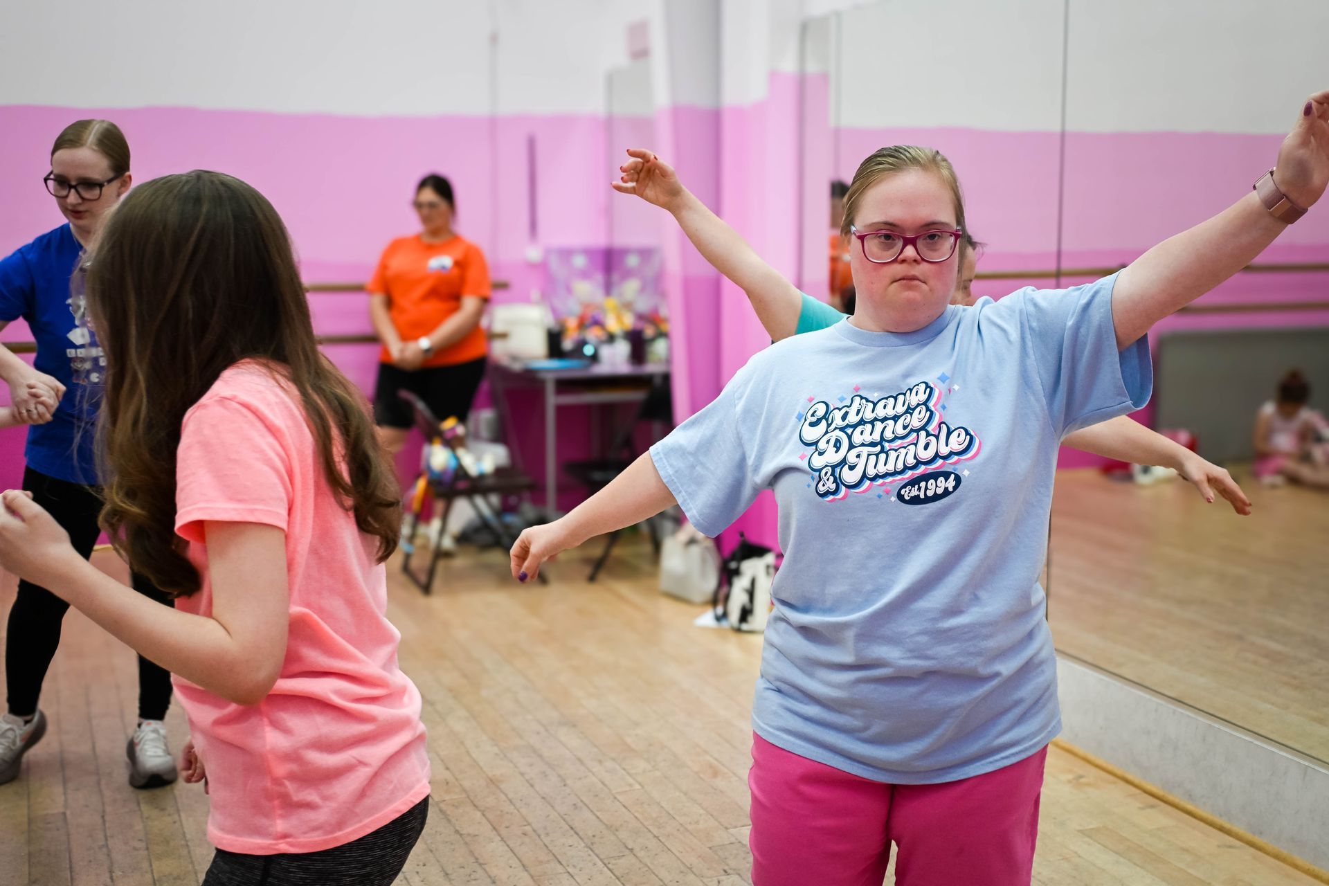 Two people dancing in a mirrored studio, one in a blue shirt and pink pants with arms raised.