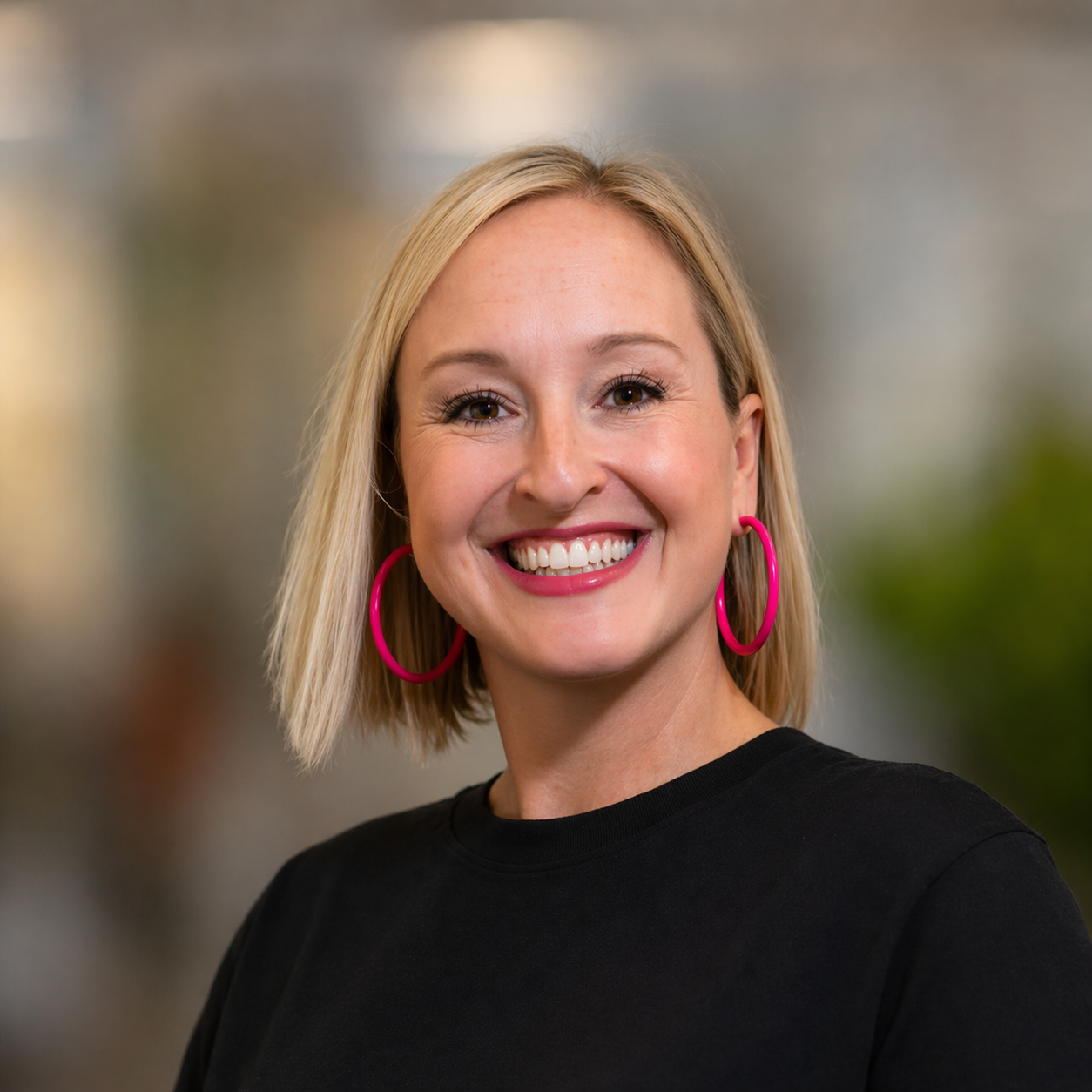 Smiling woman with blonde bob haircut and pink hoop earrings against a blurred background