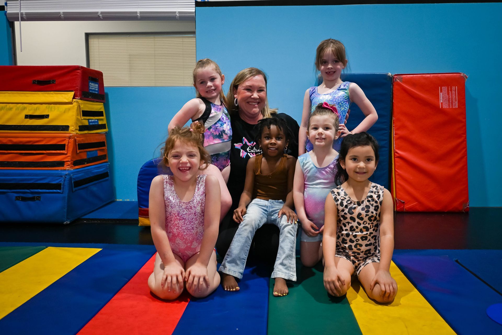 Group of children and one adult kneeling on colorful mats in a gym, smiling at the camera