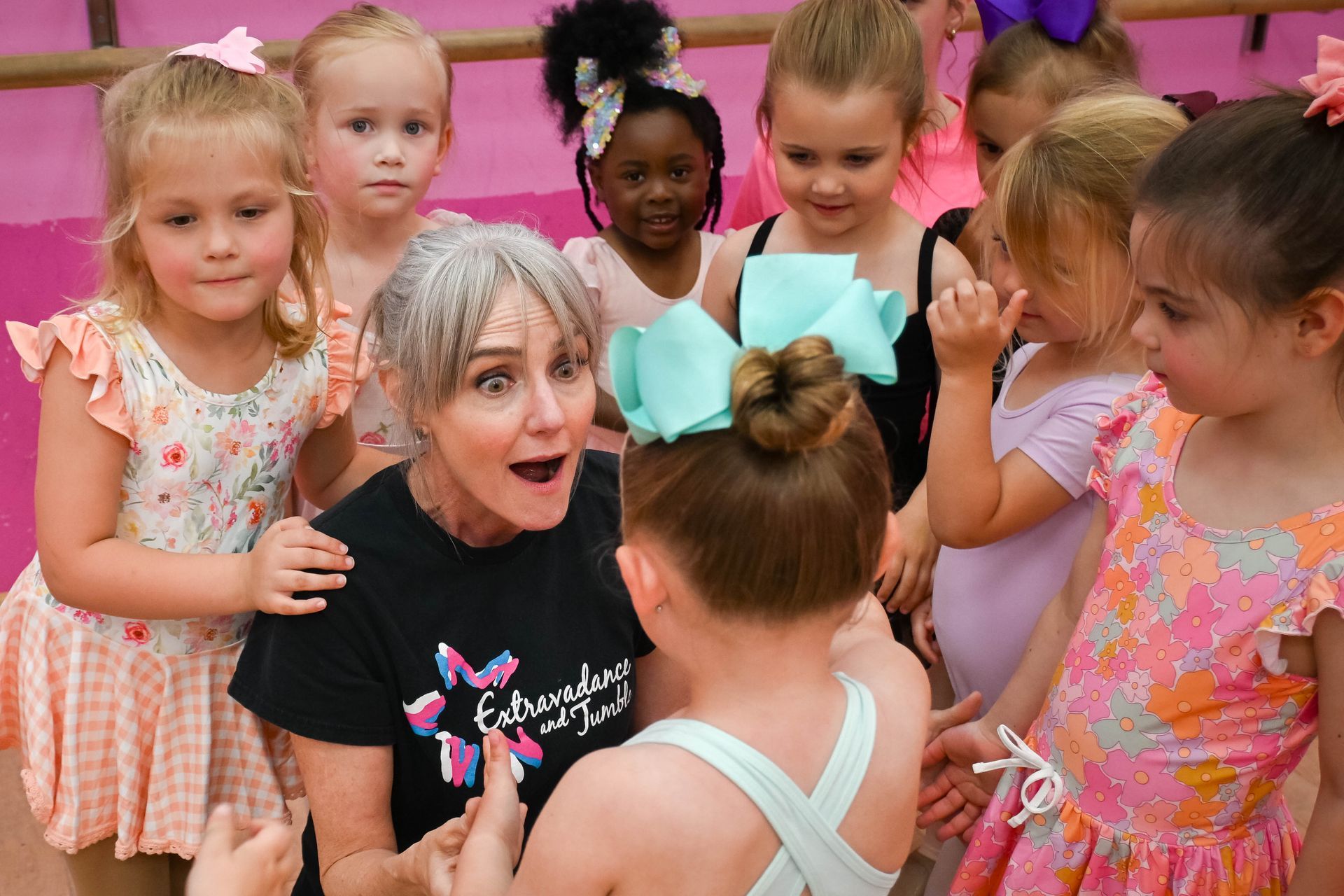 Woman surrounded by children in ballet outfits, reacting with surprise in a pink studio.