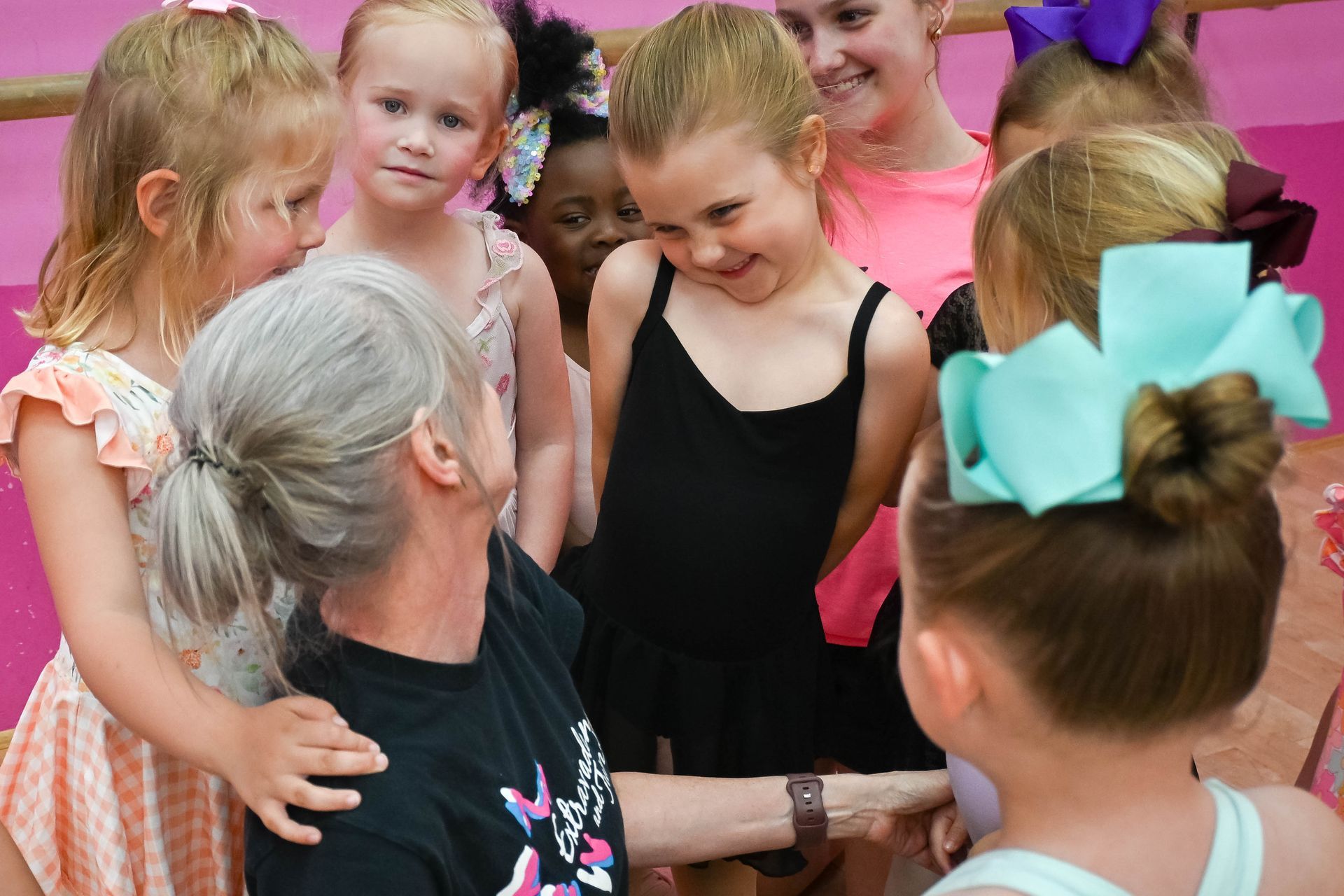 Children gather around an older woman in a lively indoor dance or class setting, smiling and chatting.