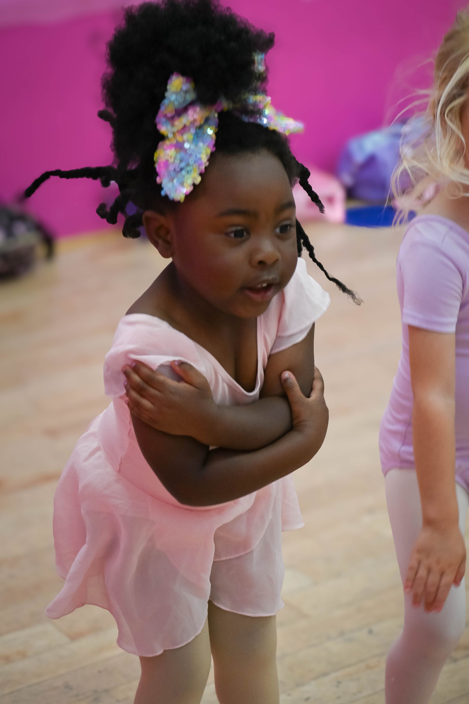 Child in a pink ballet outfit with arms crossed, dancing in a studio with a bright pink wall.