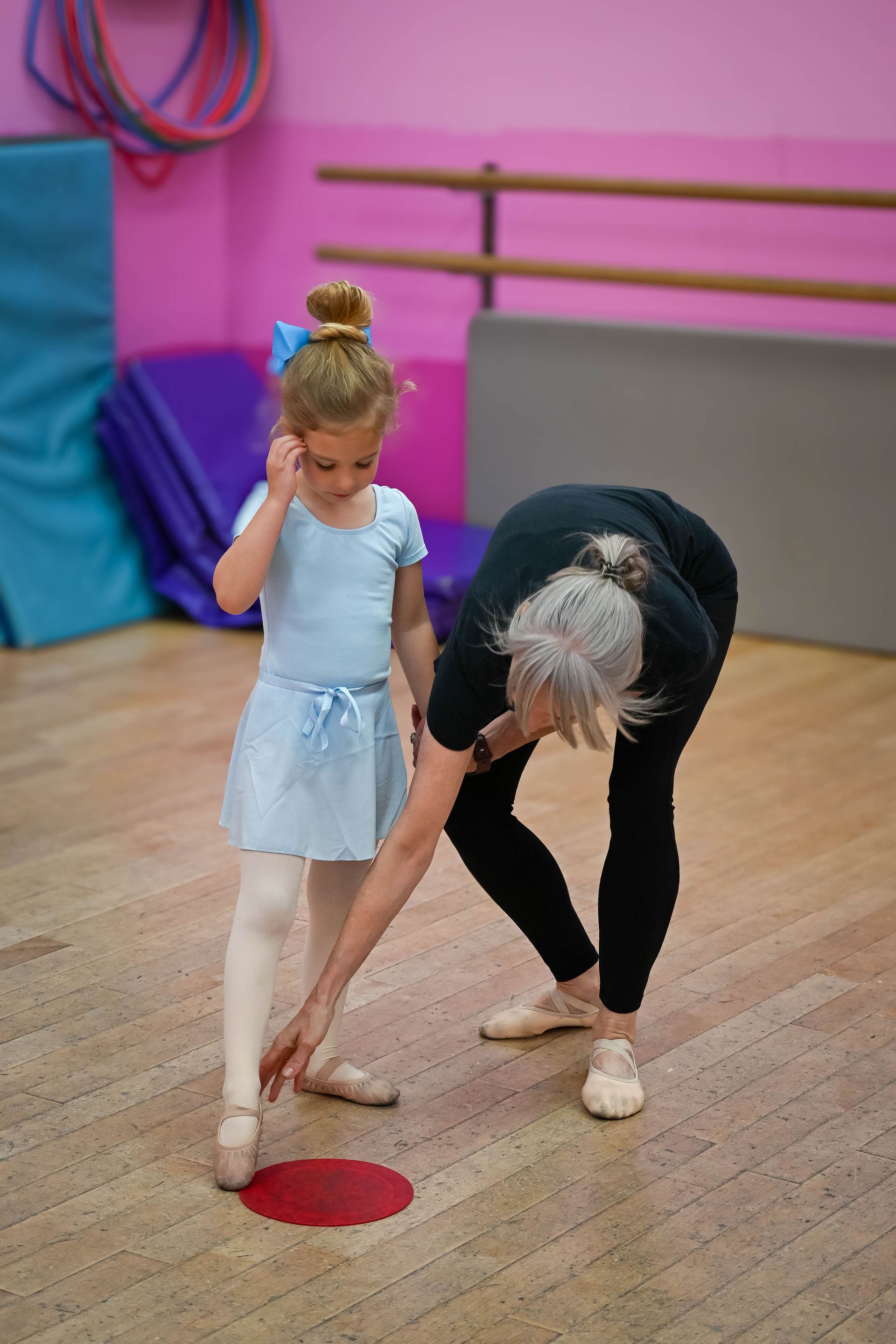 Adult bending to help a child in a dance studio with pink walls and a wooden floor