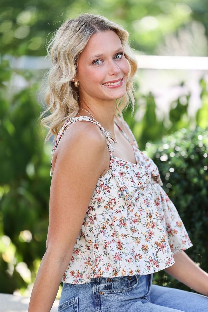 Woman smiling outdoors in a floral sleeveless top and jeans, standing in bright greenery