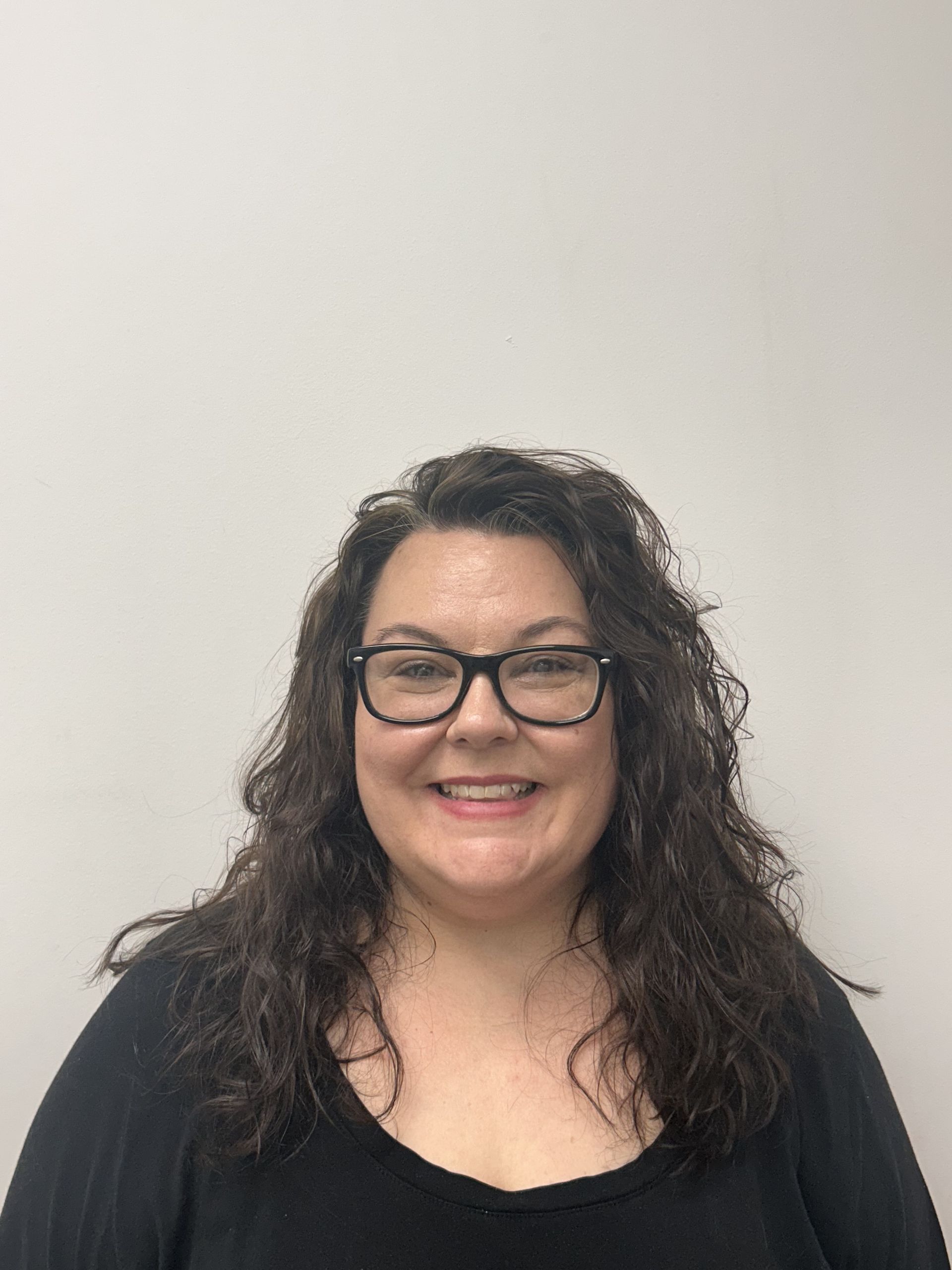 Smiling person with curly dark hair and glasses wearing a black top against a plain light background