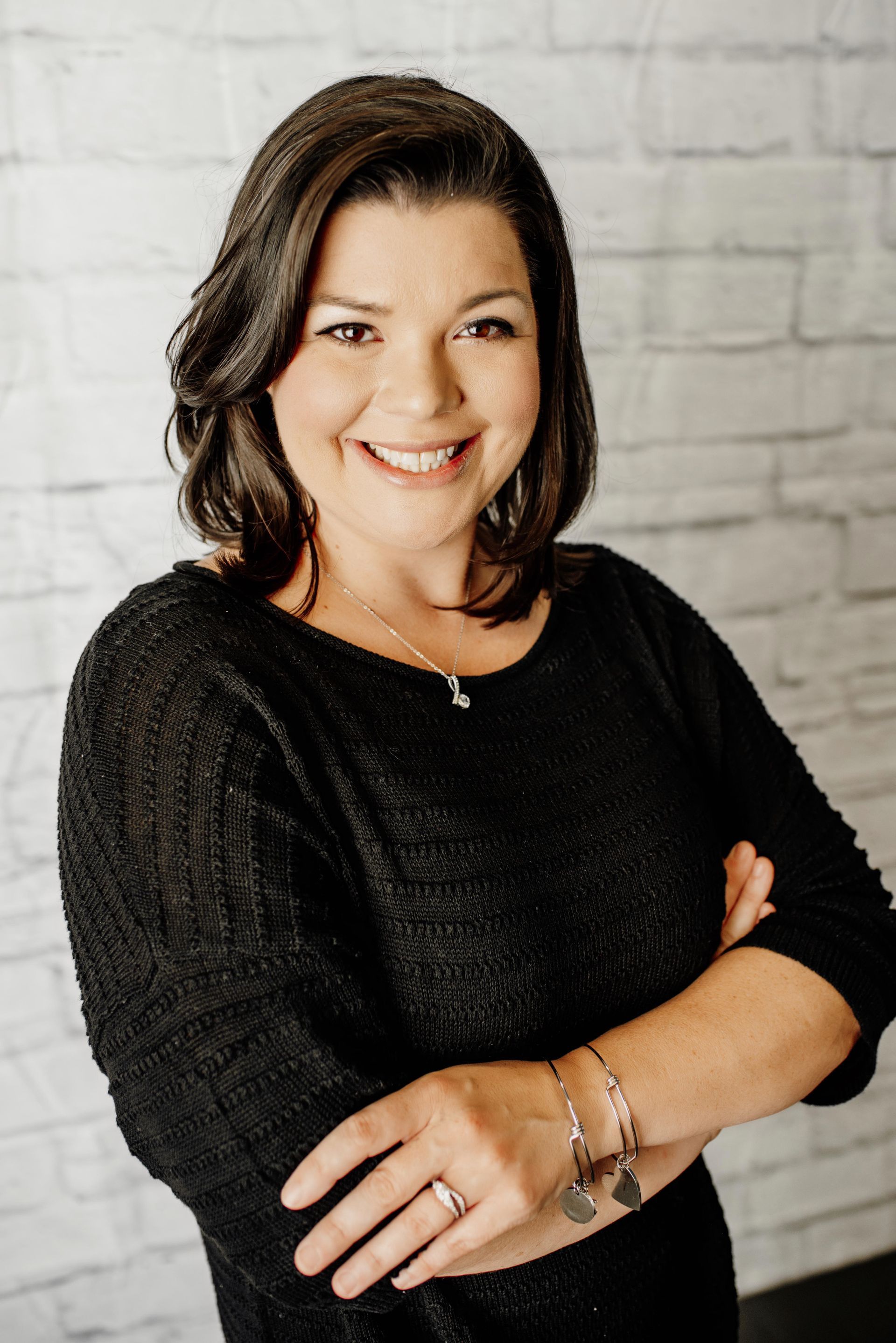 Smiling person in a black sweater with arms crossed, standing against a white brick wall.