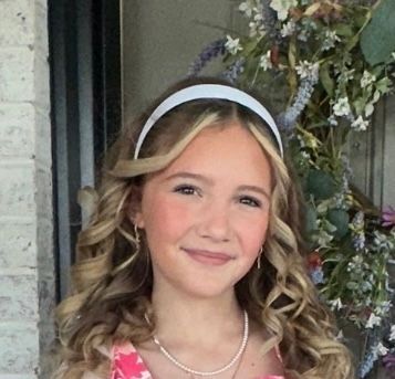 Smiling child with curly hair and white headband, standing beside a floral archway.