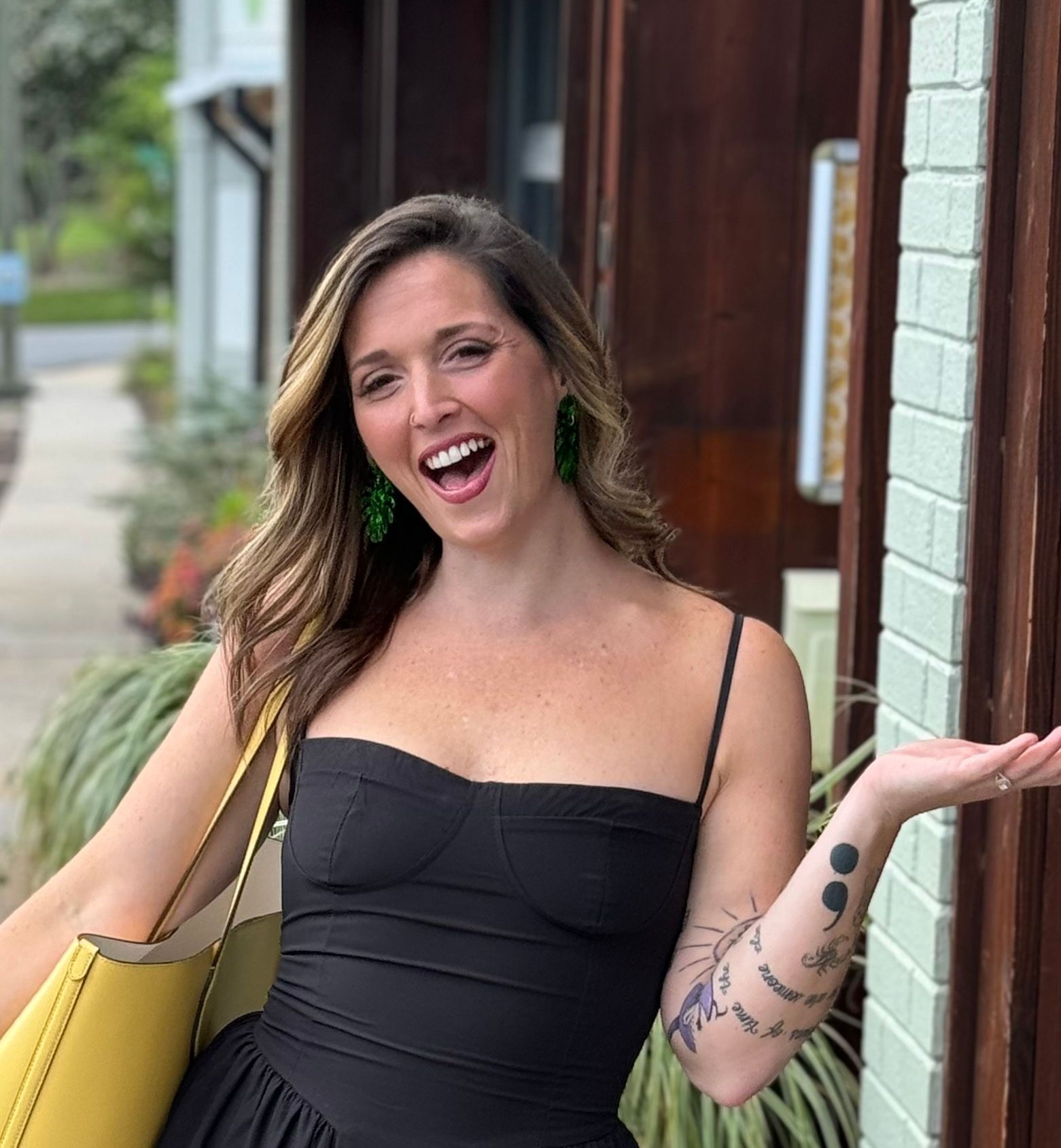 Woman smiling outdoors beside a brick wall, wearing a black dress and carrying a yellow bag.