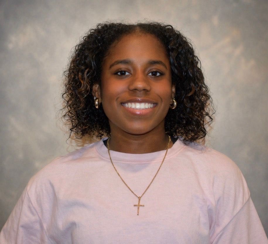 Smiling person in a light pink shirt and gold cross necklace against a gray studio backdrop
