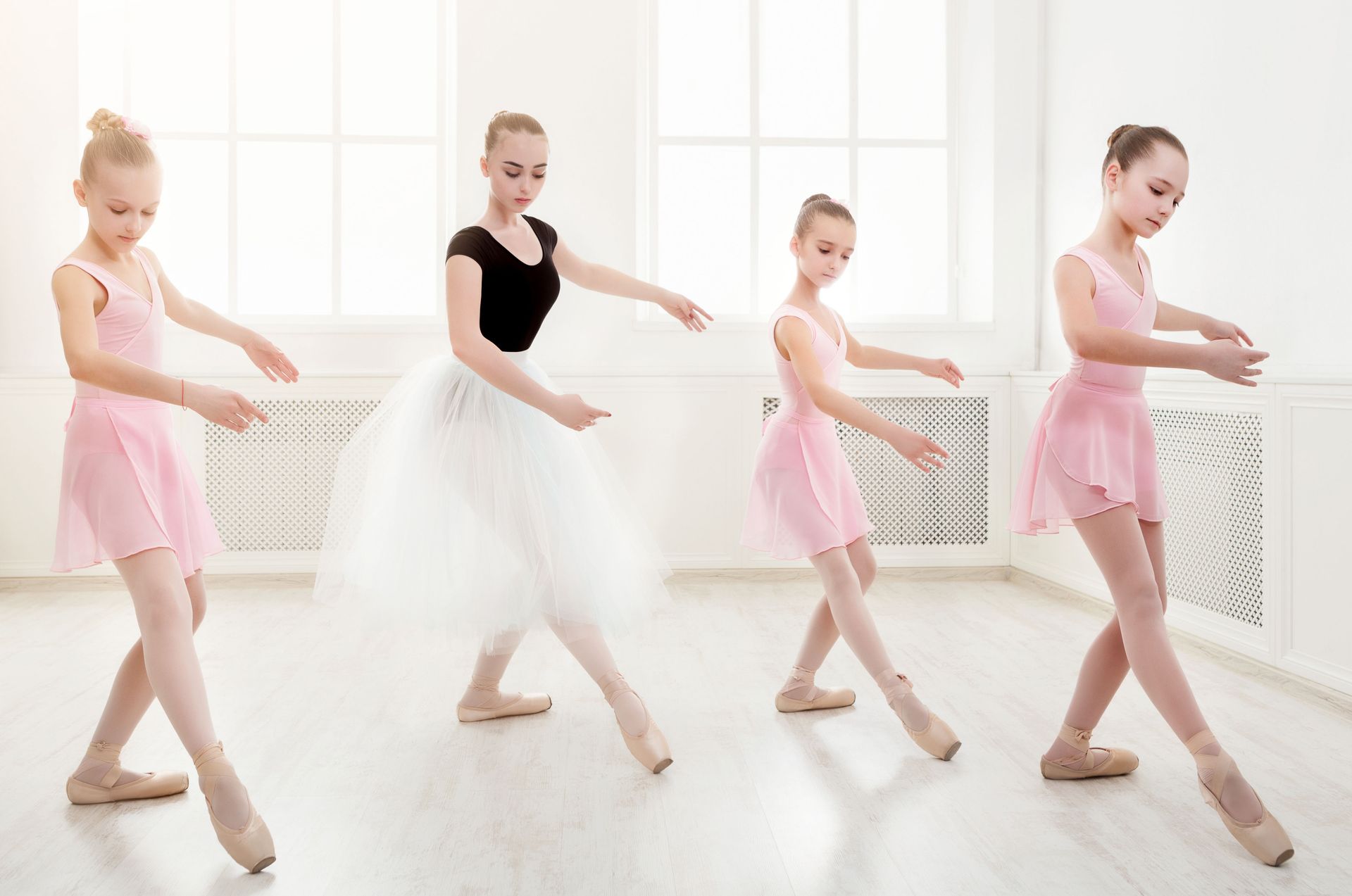 Four ballet dancers practice in a bright studio, wearing pink and white attire while performing graceful movements.