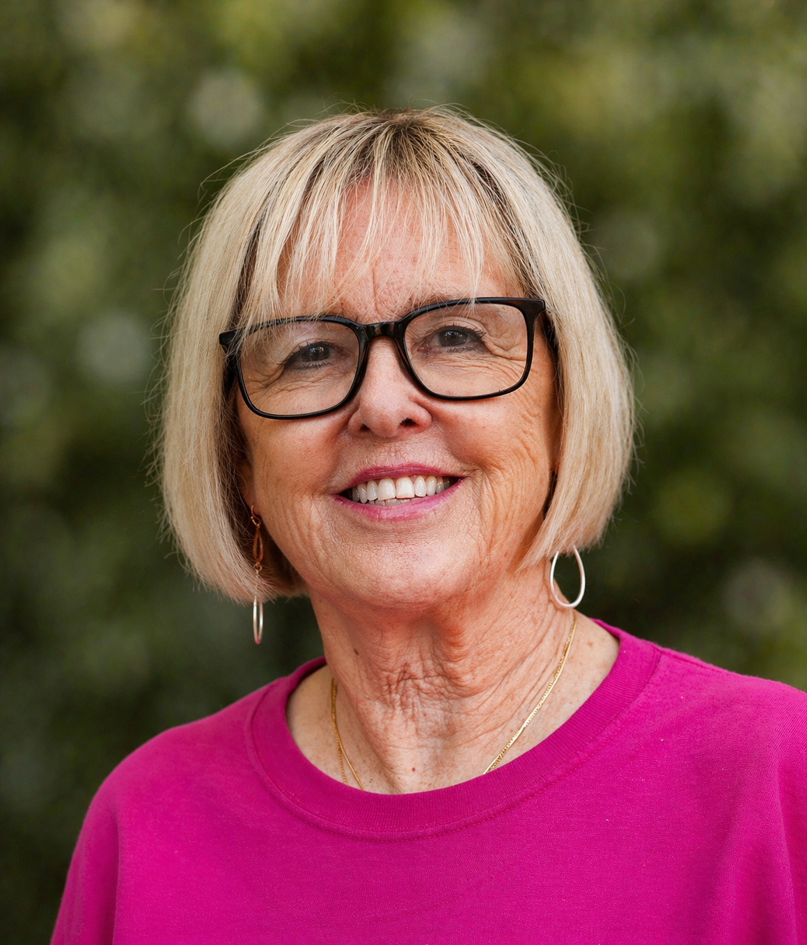 Smiling person with short blond hair and black glasses, wearing a bright pink top against a blurred outdoor background