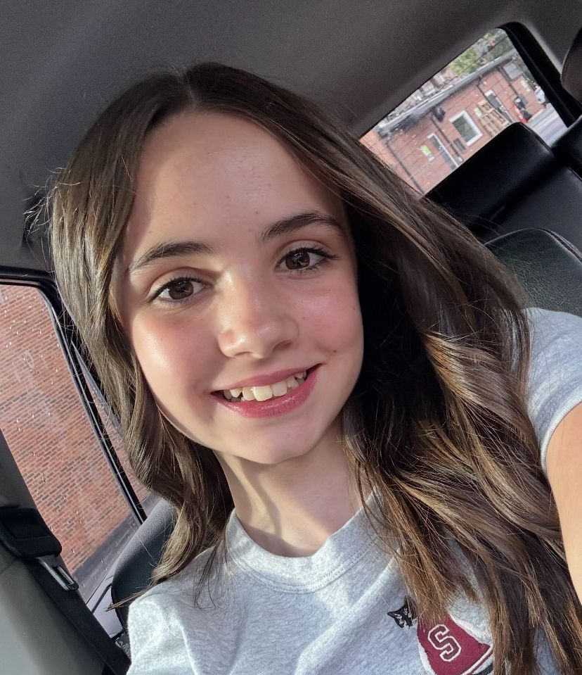 Smiling person in a car, wearing a gray shirt, with long brown hair and brick buildings outside the window