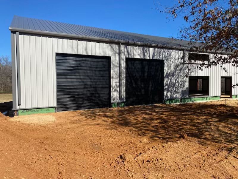 A large metal building with two black garage doors is sitting on top of a dirt field.