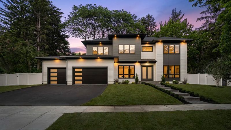 Modern two-story house with black garage doors, gray and white facade, and landscaped front yard at dusk.