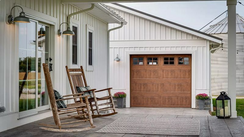 White farmhouse porch with rocking chairs, brown garage door, lantern, and potted flowers.
