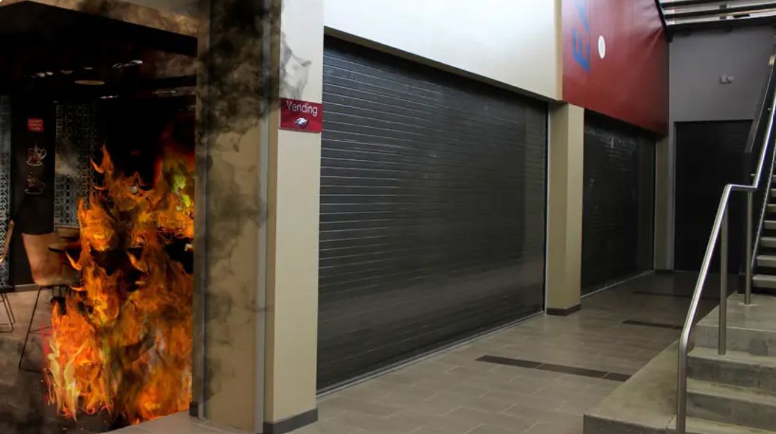 Flames engulf interior doorway; hall with closed metal security doors and stairs.
