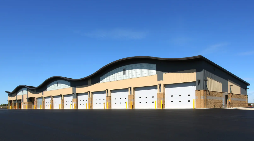 Tan industrial building with white garage doors, wavy roof, blue sky.