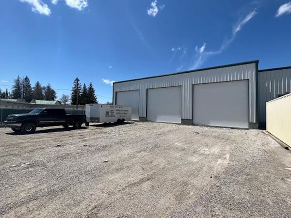 A gravel lot with a gray metal building with three closed garage doors. A black truck and white trailer are parked in front.