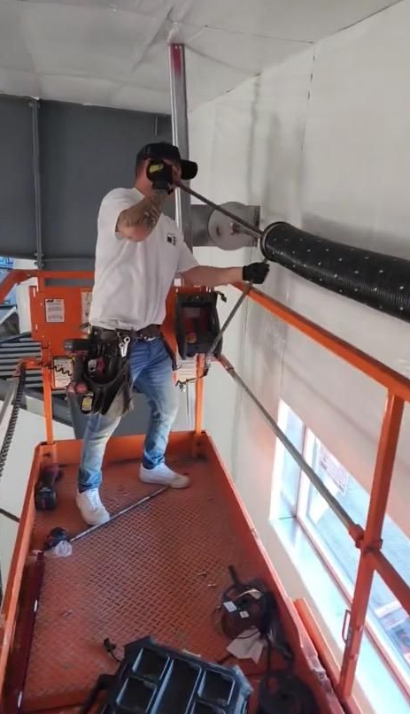 Man on lift drilling into wall, installing black roller shutter, wearing hat and safety gear.