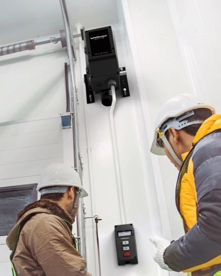 Two workers in hard hats examine a device on a white wall near a door.