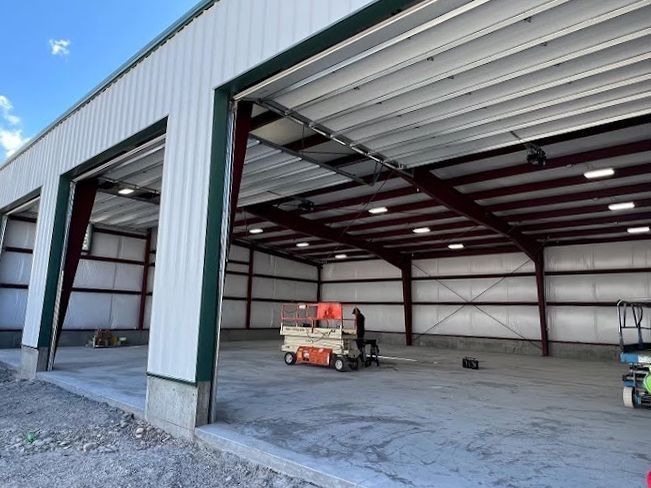 Open steel building with three bays, a person on a lift, concrete floor, red beams, and white walls.