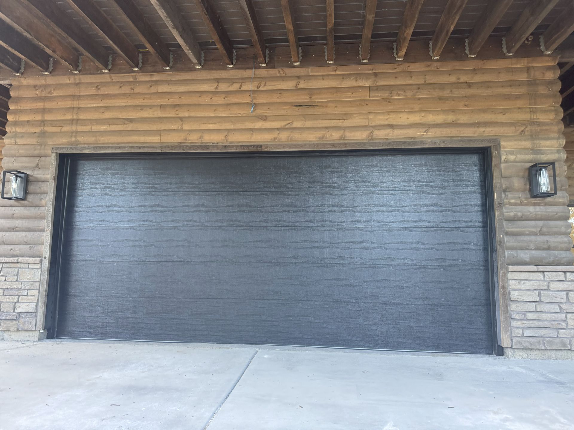Garage door with dark textured surface, flanked by stone and wood siding, under a wooden beam ceiling.