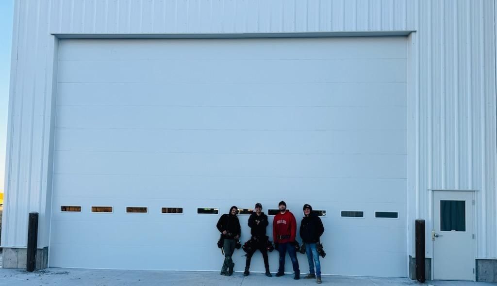 Four people stand in front of a large white garage door, building exterior.