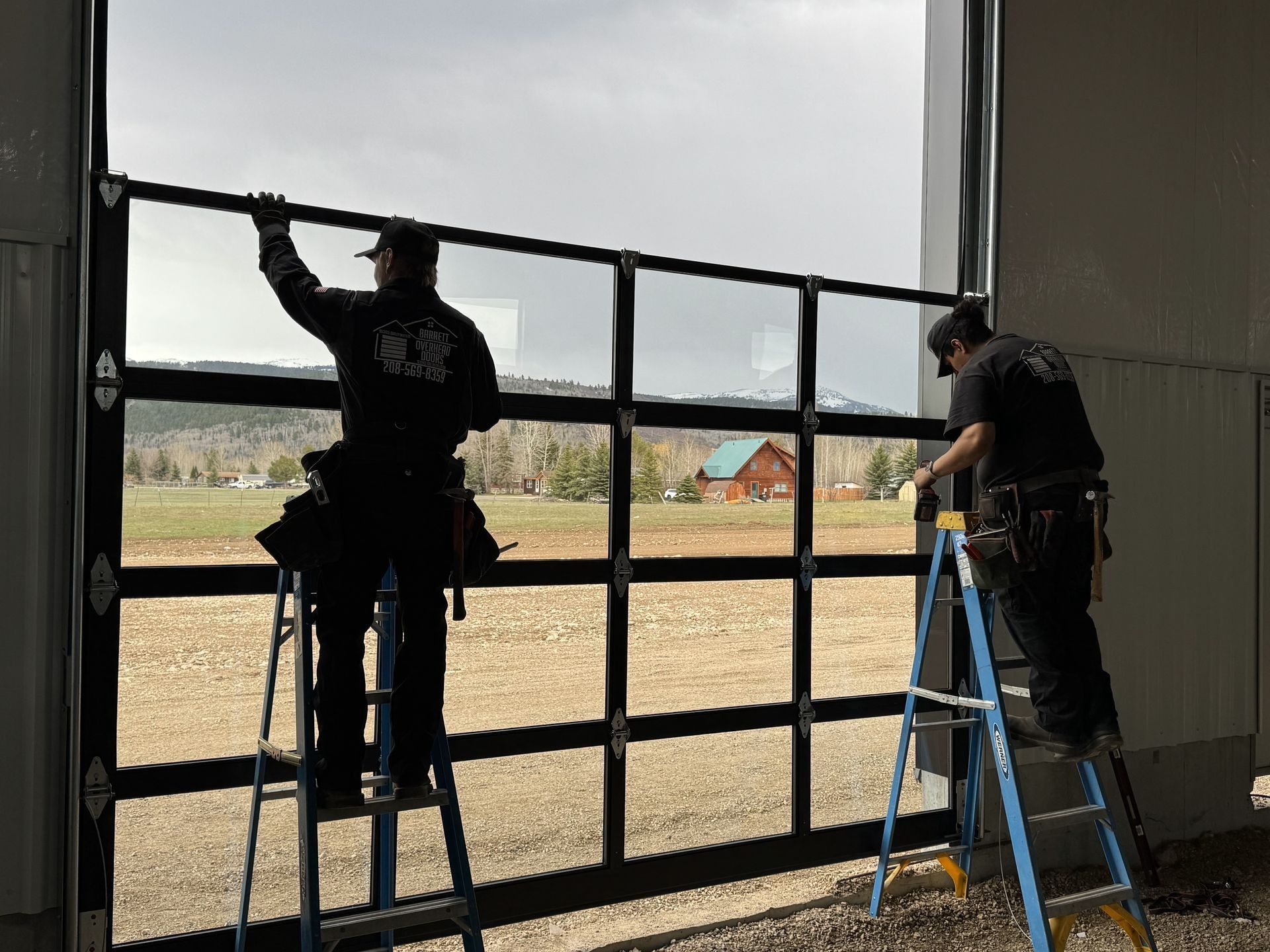 Two workers install a large glass garage door with a rural landscape in the background.