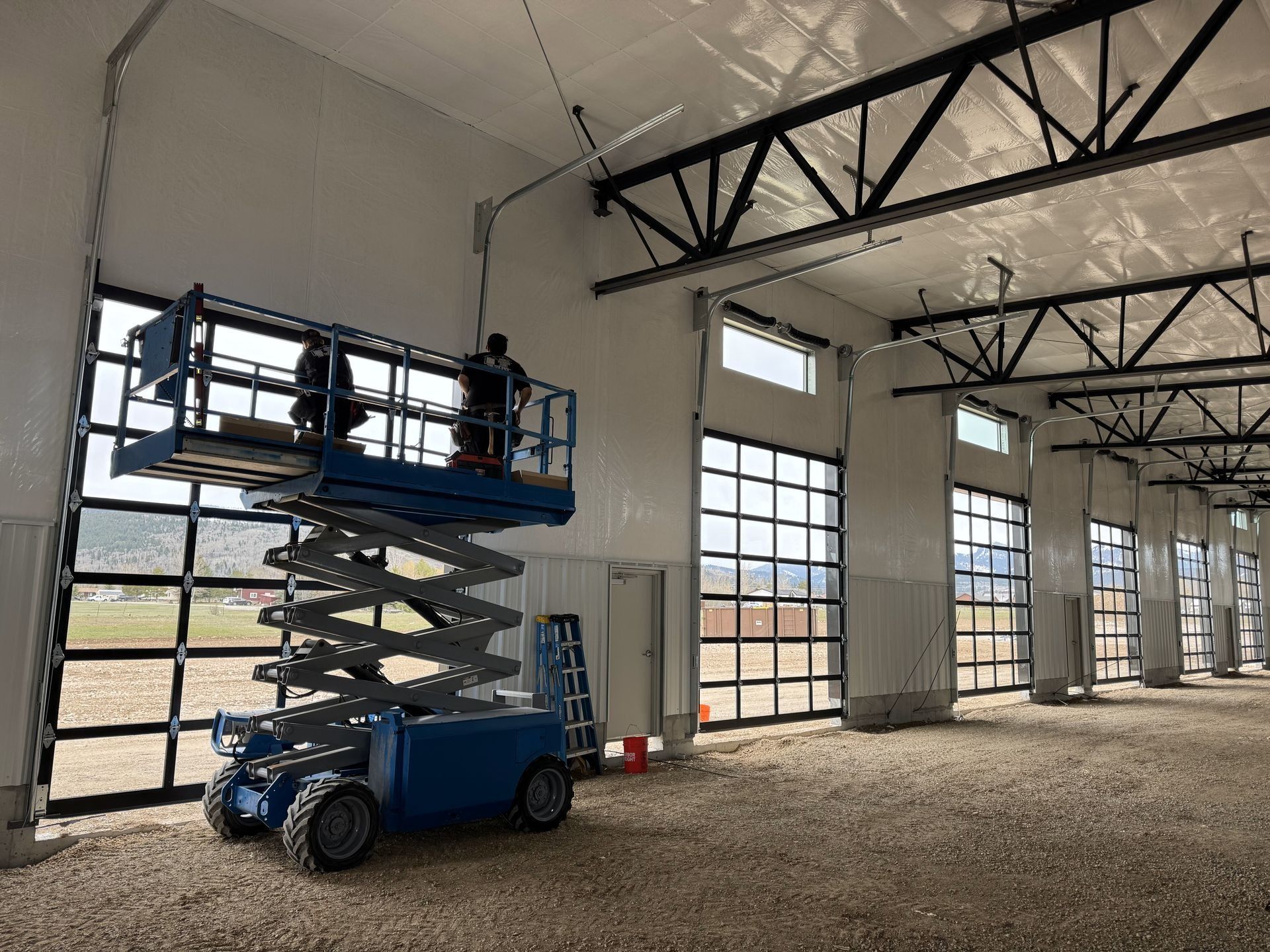 Workers on a blue lift installing wiring near the ceiling of a large building with glass doors.