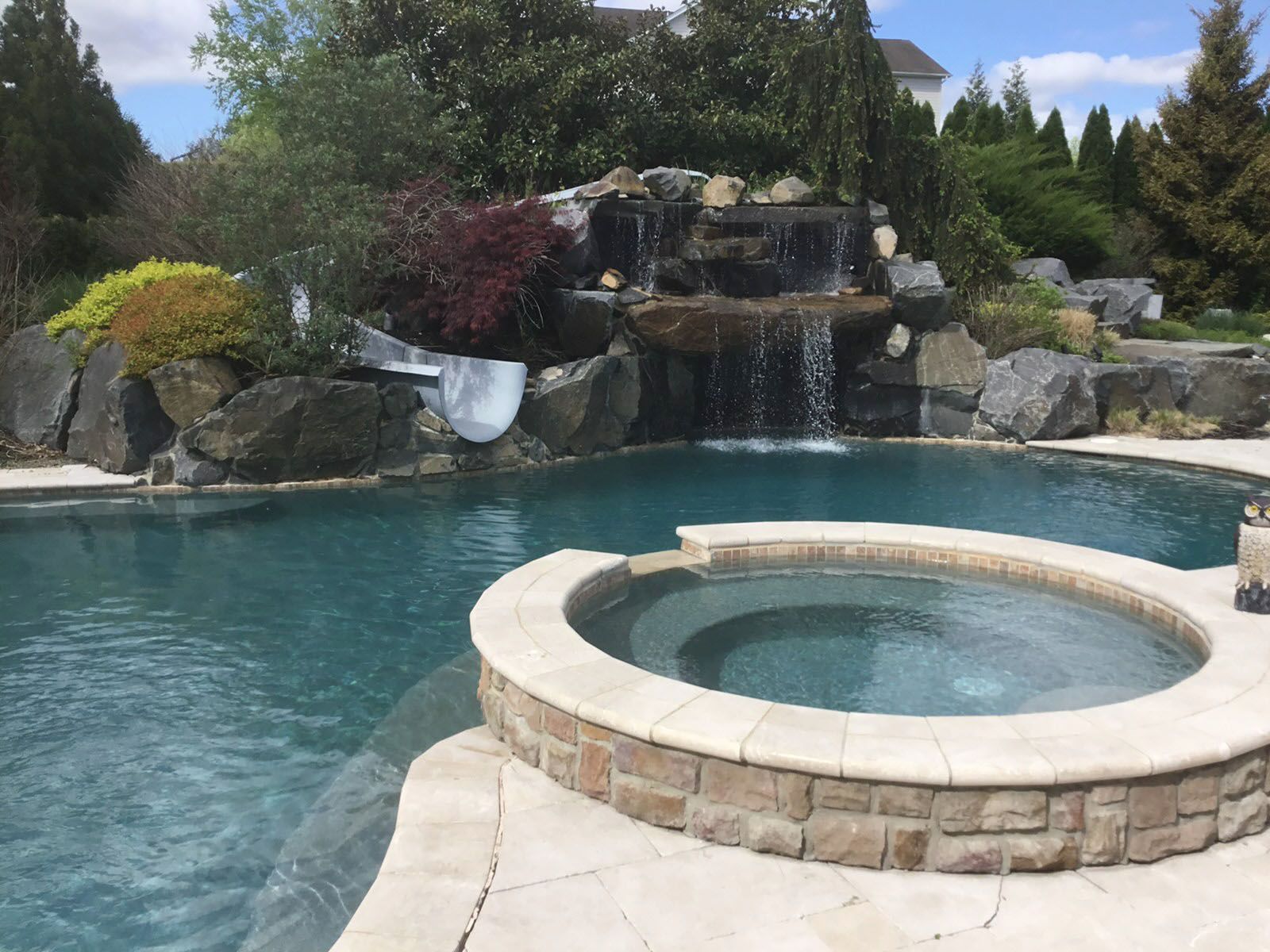 Backyard pool with waterfall, slide, and a hot tub, blue water, and stone features