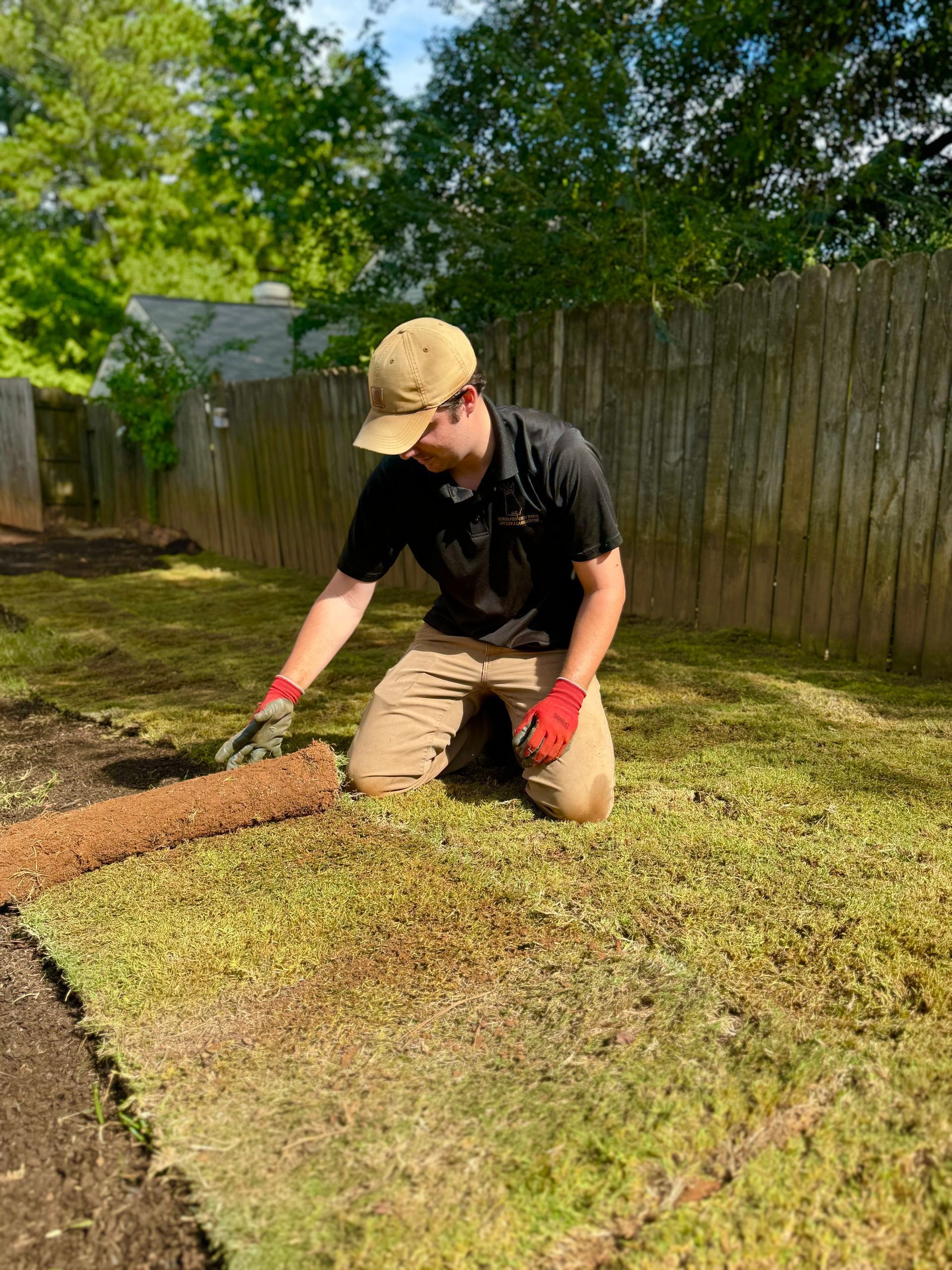 Man kneeling, rolling out sod in a backyard, wearing a hat, black shirt, and khaki pants.
