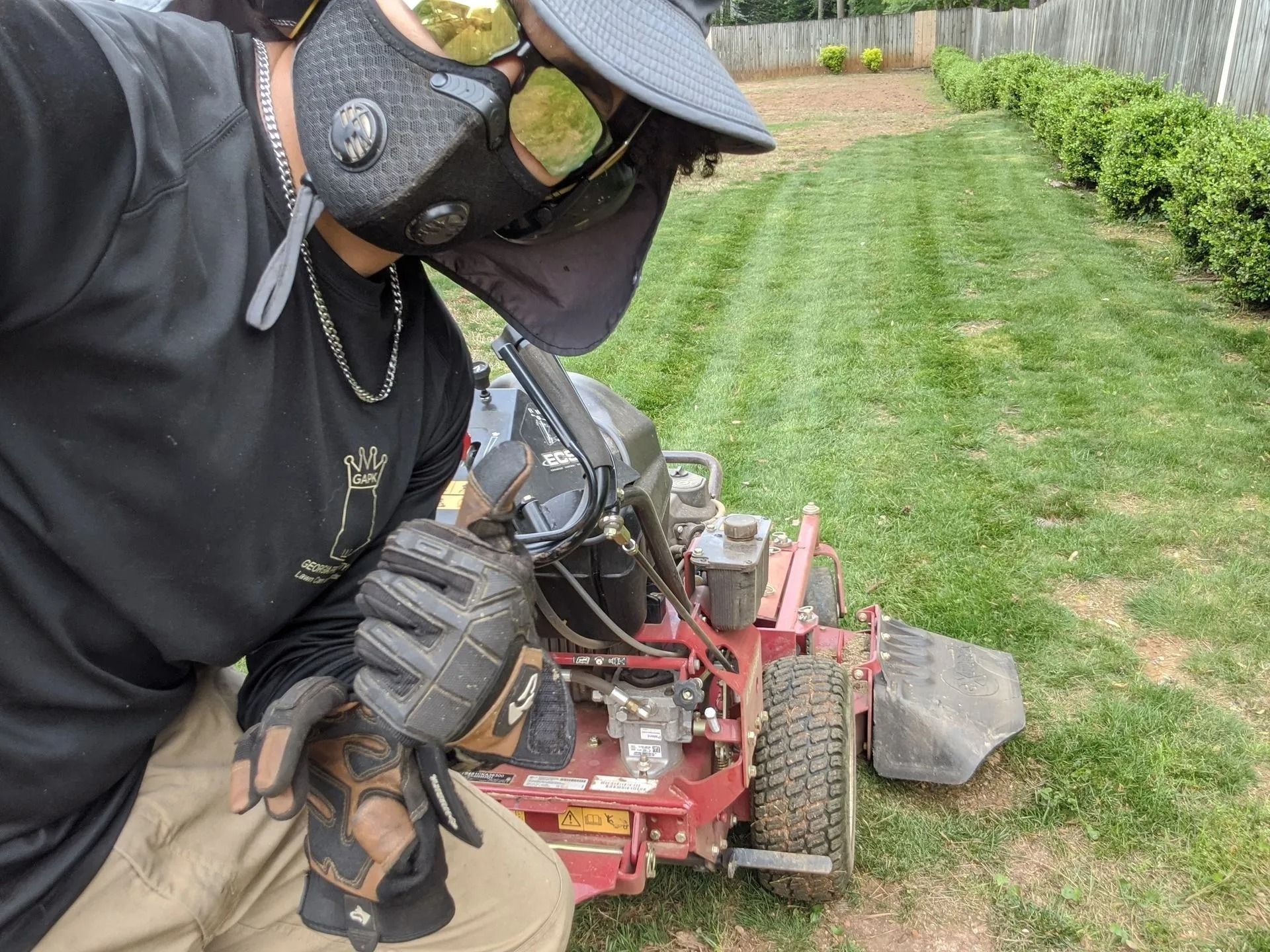 Person operating a red lawnmower on a green lawn. Wearing a protective mask, sunglasses, and gloves.