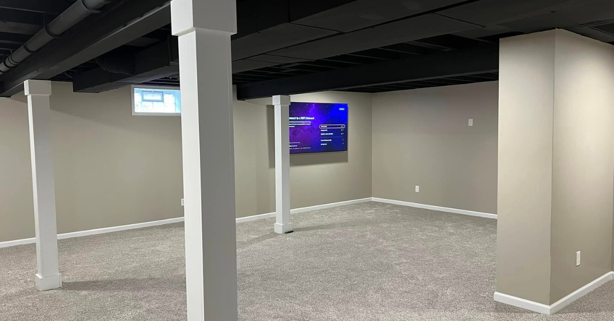 Empty basement room with gray walls, carpet, white columns, and two wall-mounted screens.