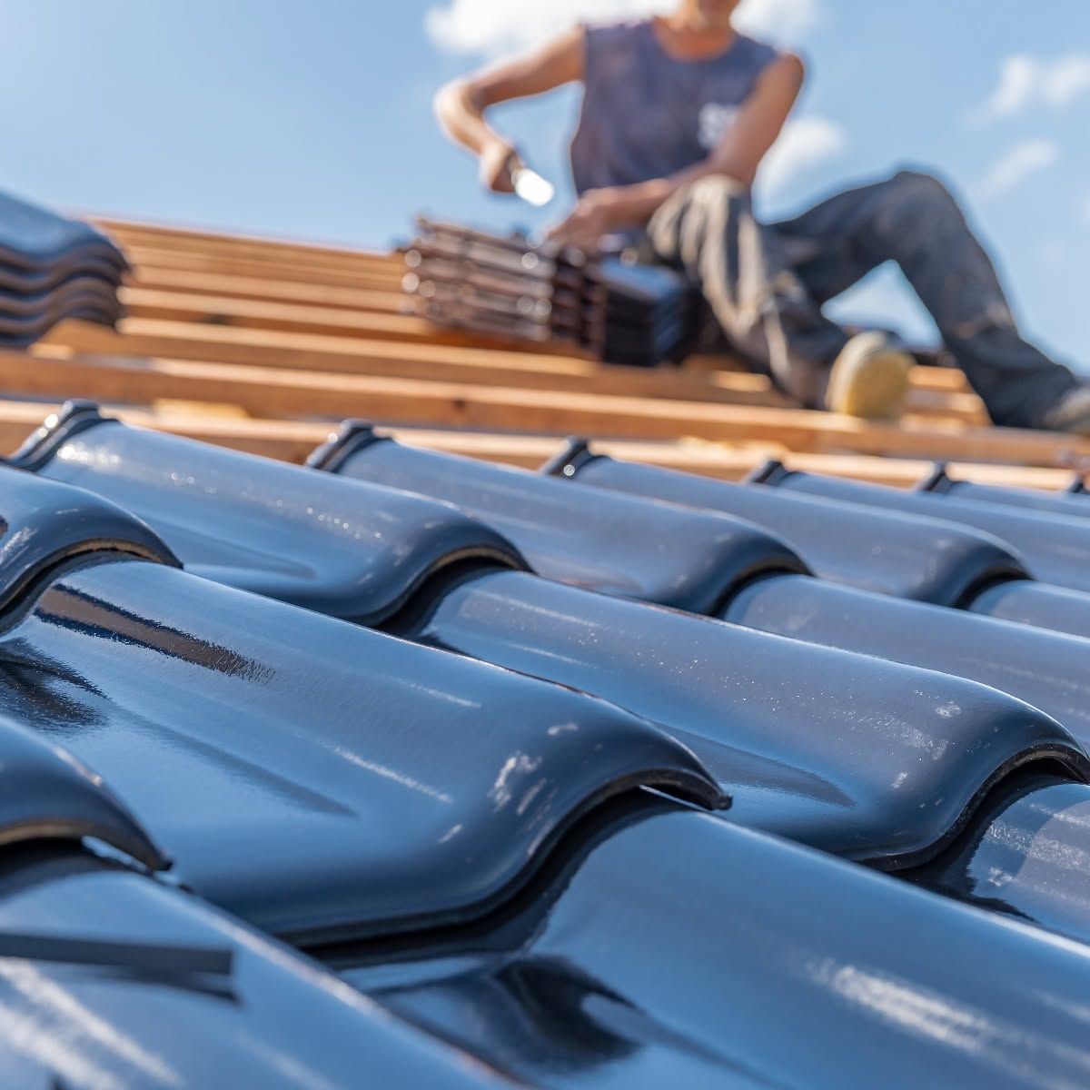 Blue roof tiles in foreground with a blurred worker installing wooden rafters in the background
