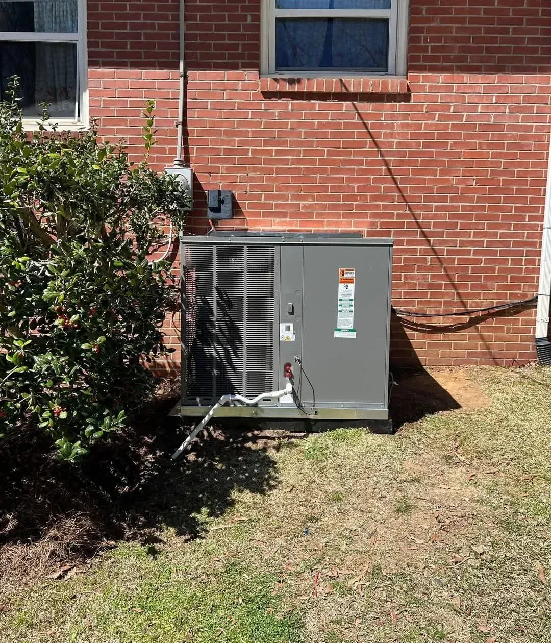 Gray outdoor HVAC unit beside a brick house wall, partly obscured by shrubs and grass