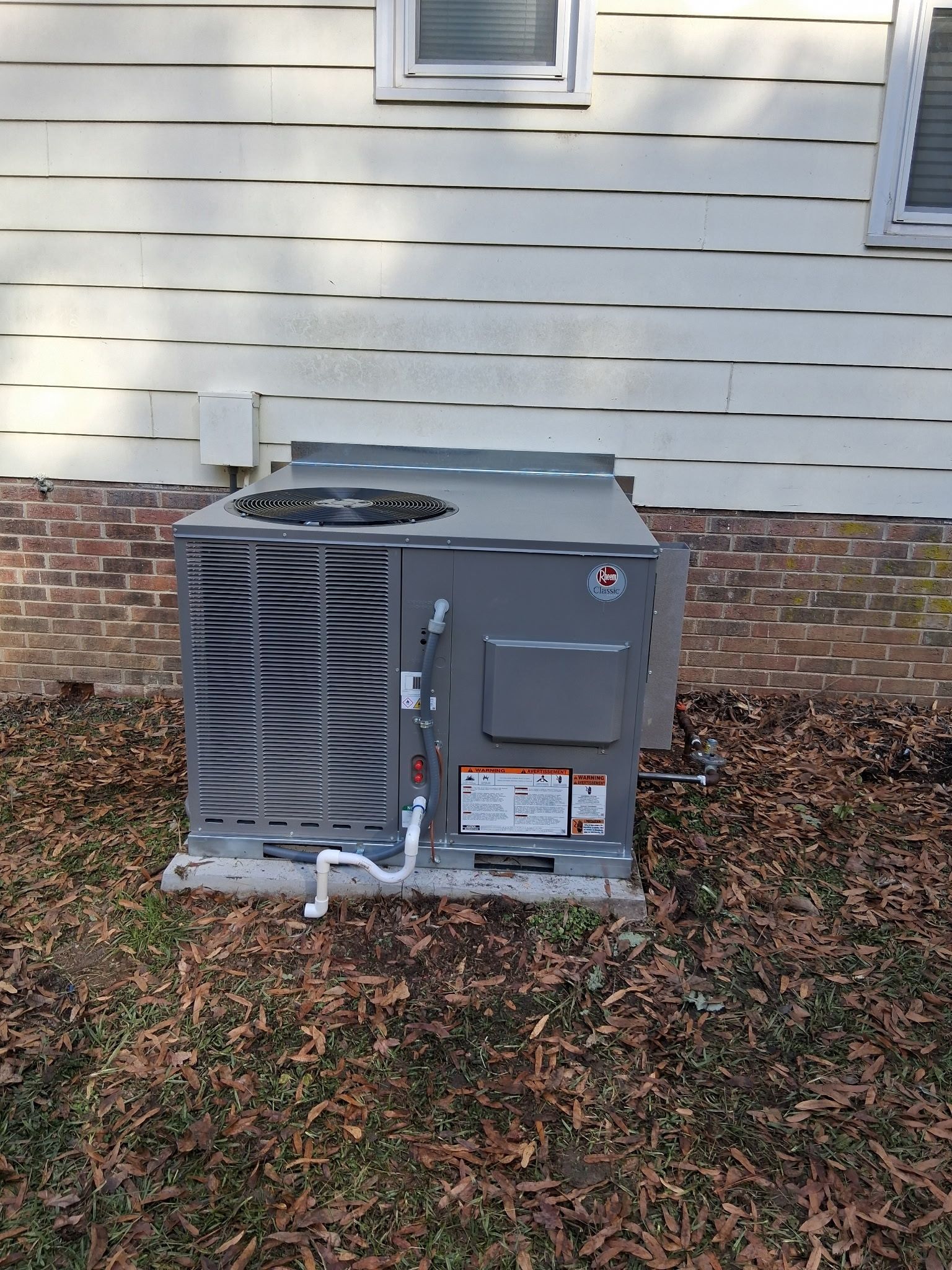 Outdoor HVAC unit beside a house with white siding and a brick foundation, sitting in a leaf-covered yard