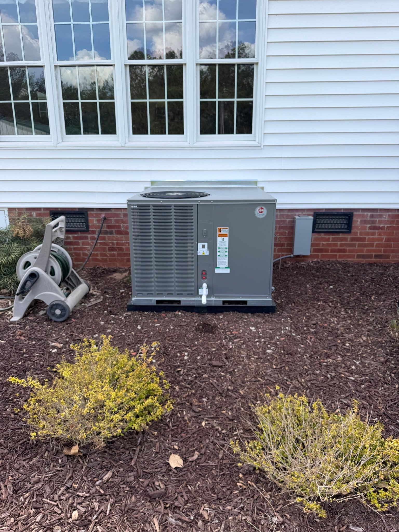 Outdoor air conditioning unit beside a white house wall and window, with landscaping shrubs in front.