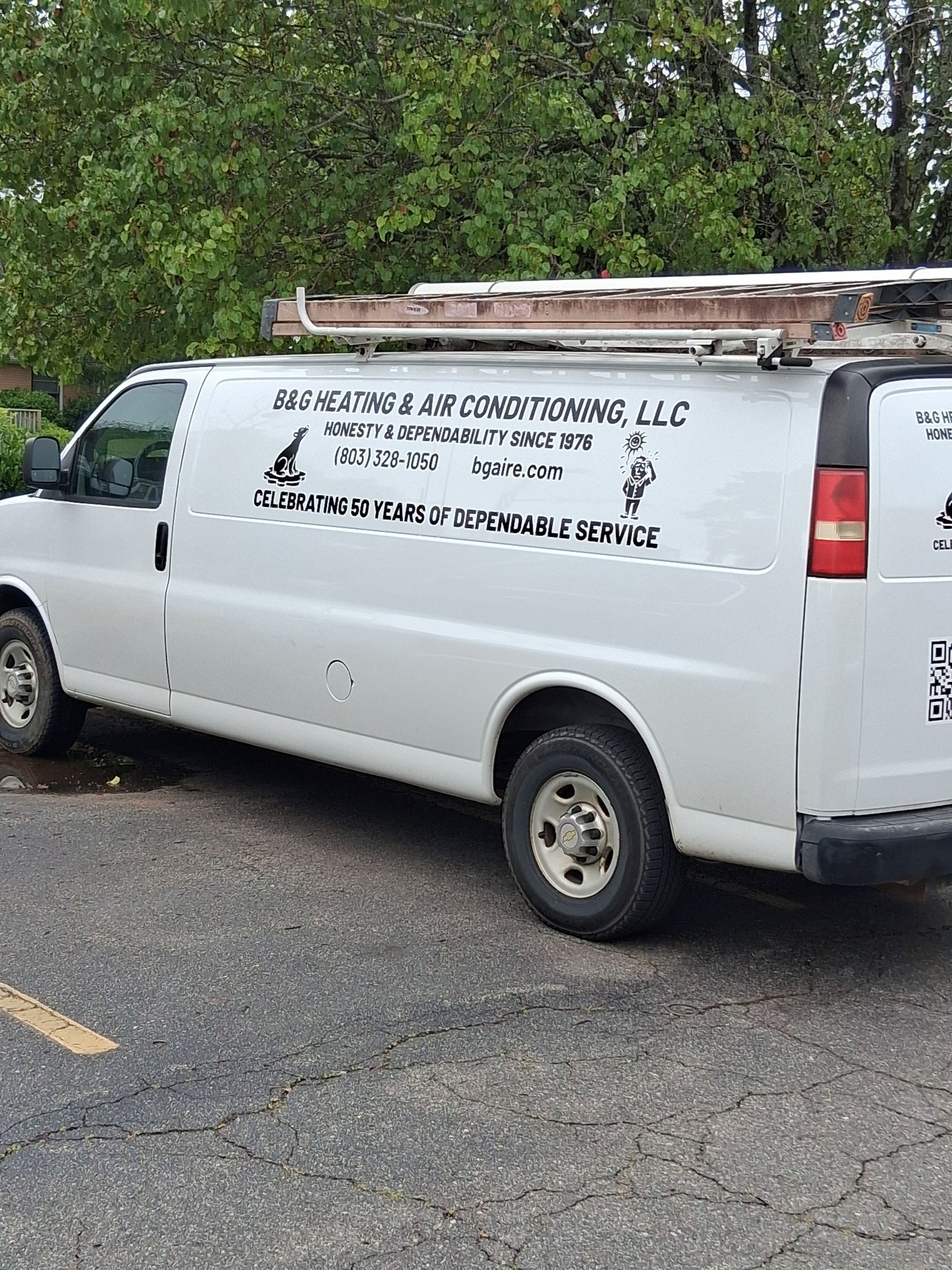 White work van with ladder rack and company lettering parked on a road beside trees.