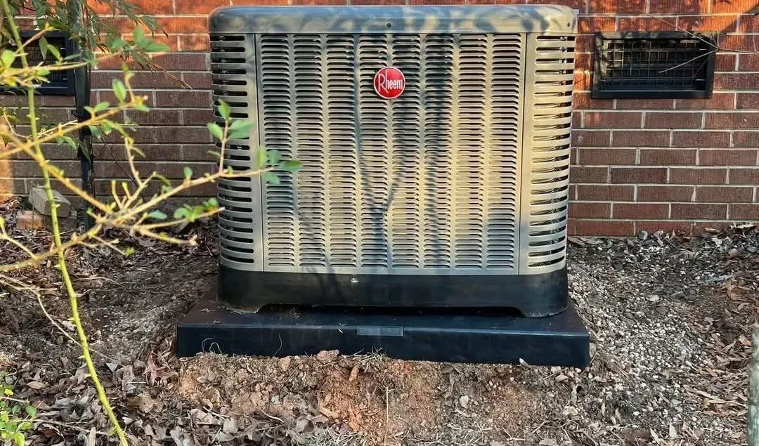 Outdoor air conditioner unit beside a brick wall, with a red sticker and gravel landscaping around it.