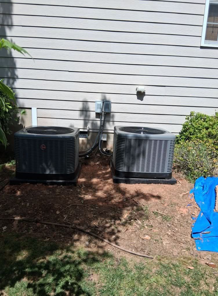 Two outdoor HVAC units beside a house wall, one black and one gray, with hoses between them.