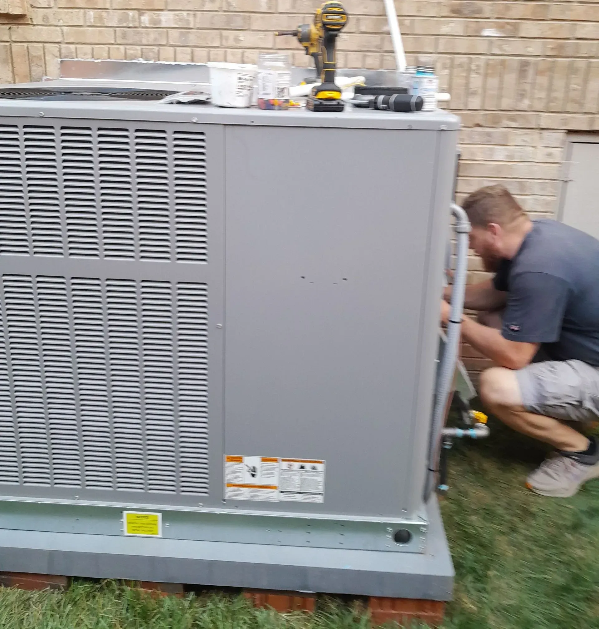Technician kneels beside a large gray outdoor HVAC unit against a brick wall.